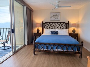 A bedroom with a bed, two bedside lamps, a ceiling fan, a framed picture, and a view of the ocean through a sliding door. - Oceanfront Whale Watching Sunsets Island Views on Big Lanai (Lahaina)