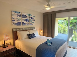 A hotel room with a bed, a ceiling fan, artwork, a nightstand, a lamp, and a view of greenery through a window.