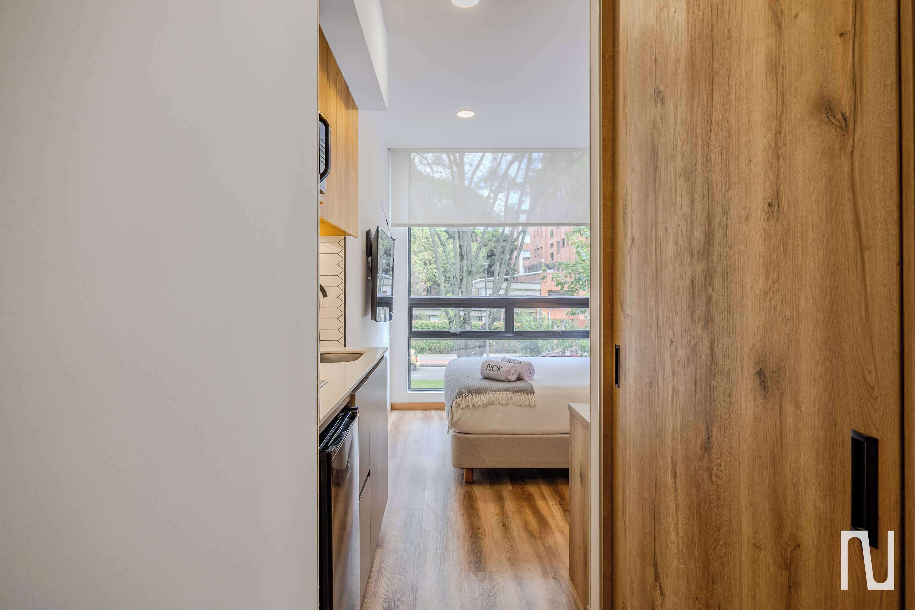A modern hotel room with a wooden floor, a bed with a white headboard, a window with a view of trees and buildings, and a kitchen area with a sink and cabinets.