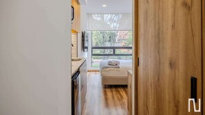 Une chambre d’hôtel moderne avec un plancher en bois, un lit doté d’une tête de lit blanche, une fenêtre donnant sur des arbres et des immeubles, et un coin cuisine équipé d’un évier et de placards.