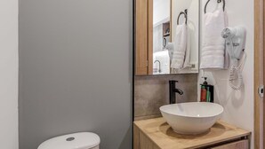Une salle de bain moderne avec des toilettes blanches, un meuble-lavabo en bois doté d’un grand évier, un miroir et un sèche-cheveux fixé au mur.