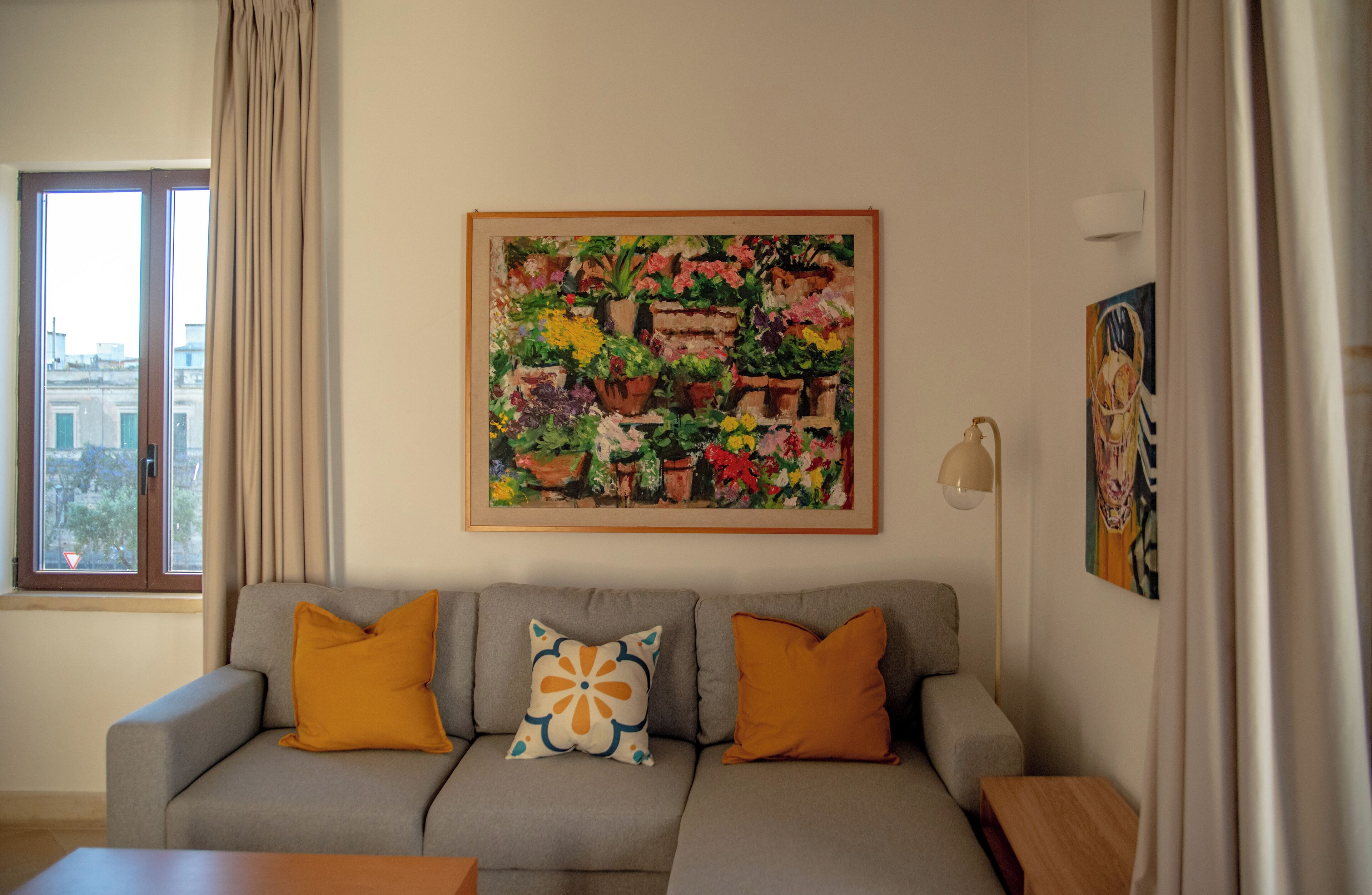 Un salon avec un canapé gris, deux coussins orange, un tableau floral encadré et une lampe.