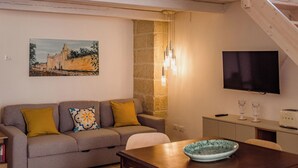 Un salon chaleureux avec un plafond haut, un escalier en bois, un canapé avec des coussins, une table à manger avec des chaises, un téléviseur et un tableau accroché au mur.
