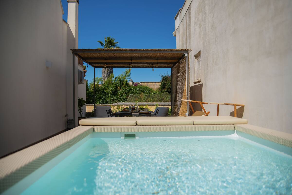 A swimming pool with a sunlit patio area, outdoor seating, and a view of surrounding greenery.