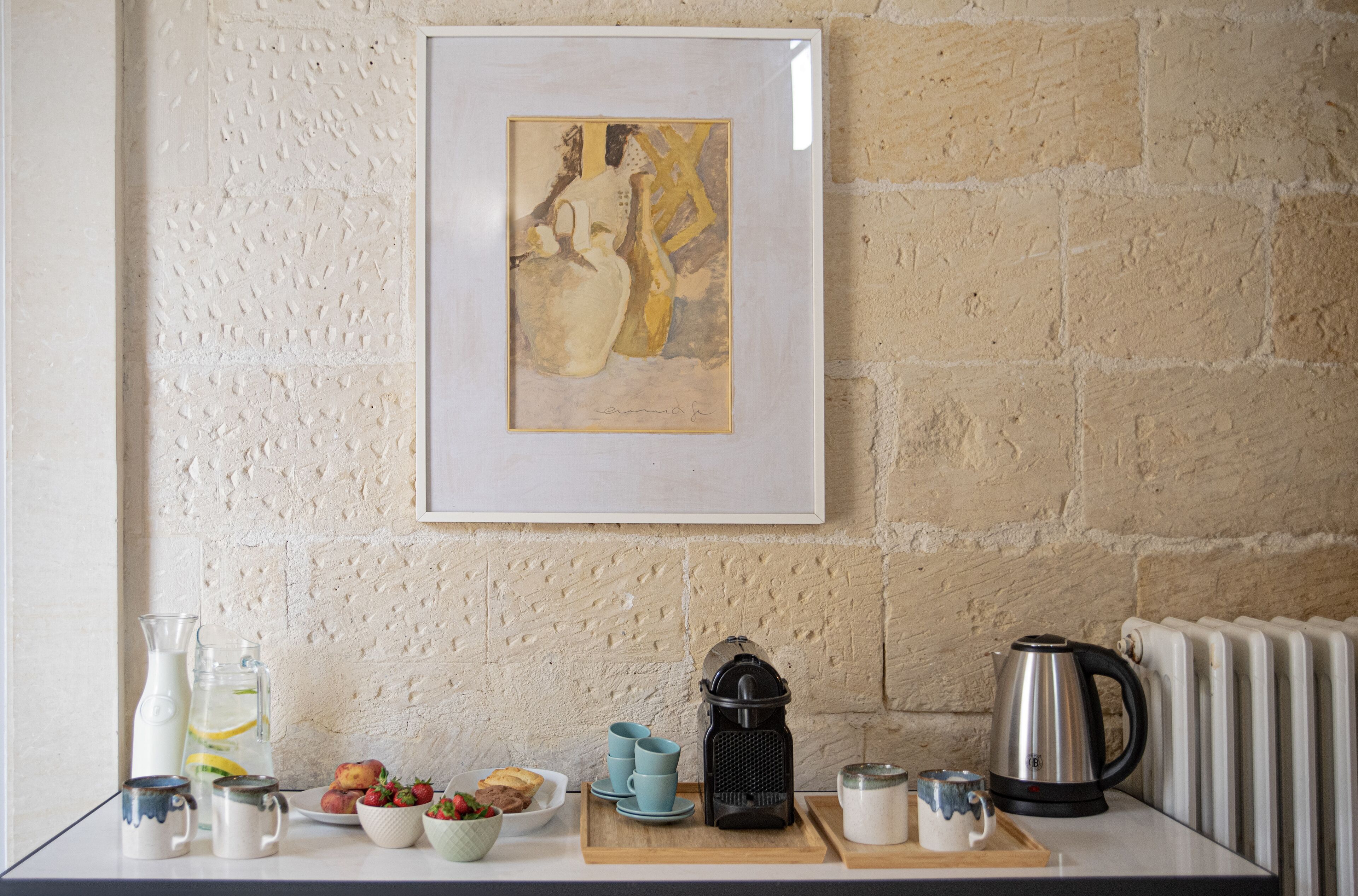 Uma cozinha com parede de pedra, uma obra de arte emoldurada e diversos utensílios, como uma chaleira, xícaras e uma fruteira.