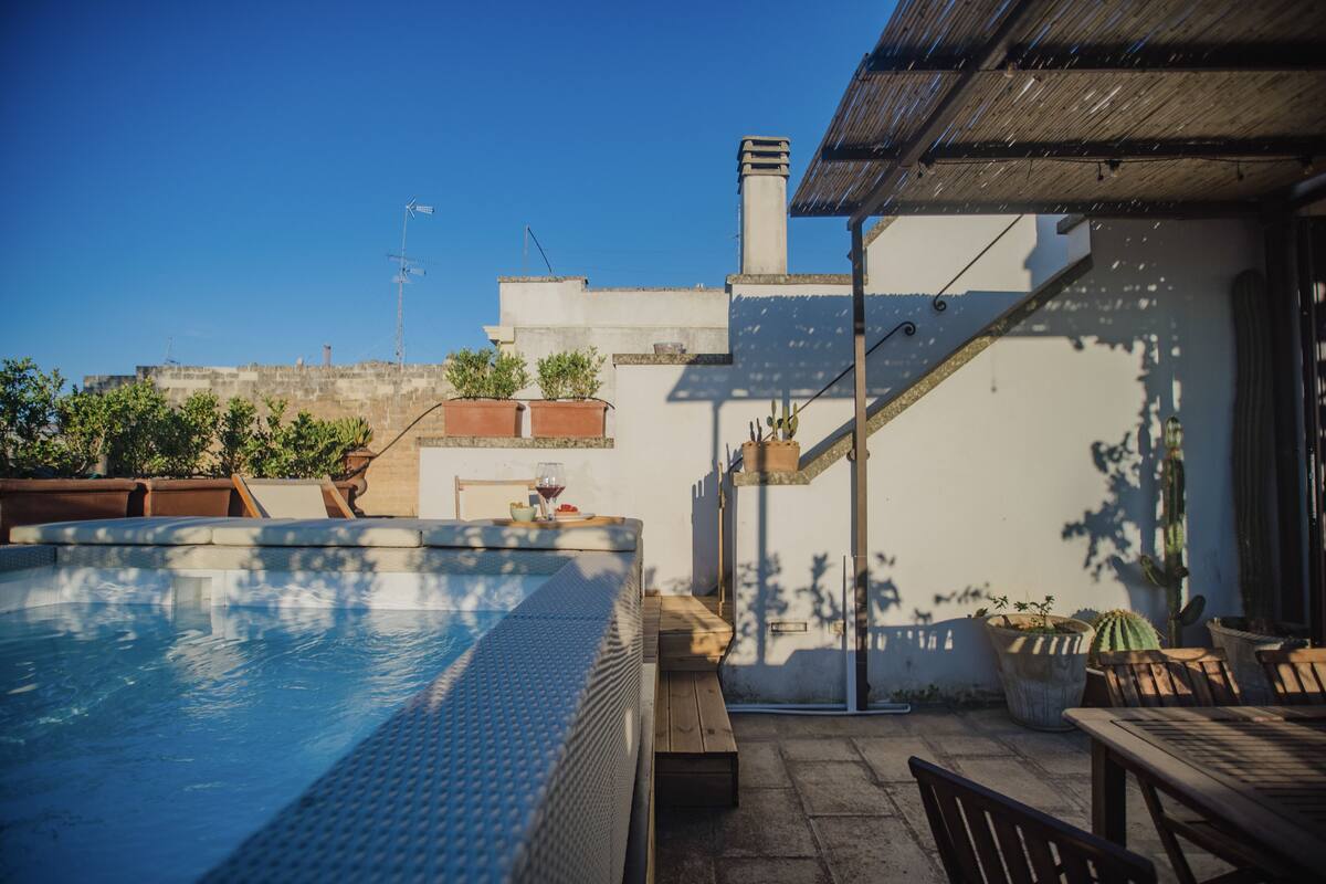 A rooftop swimming pool with a wooden deck and seating area, surrounded by potted plants and a clear view of the city skyline.