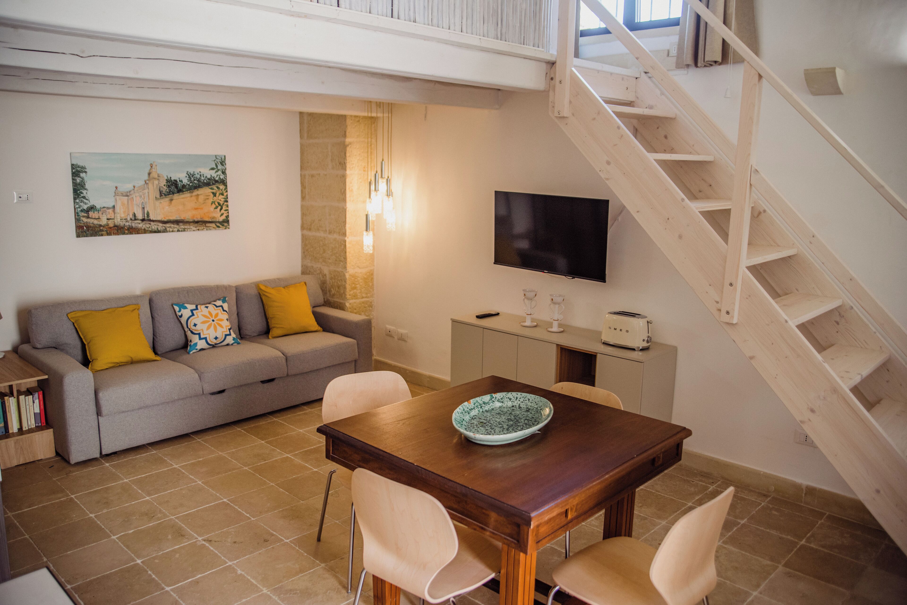 Una sala de estar con un sofá, una mesa de comedor con sillas, un televisor, una escalera y un cuadro en la pared.