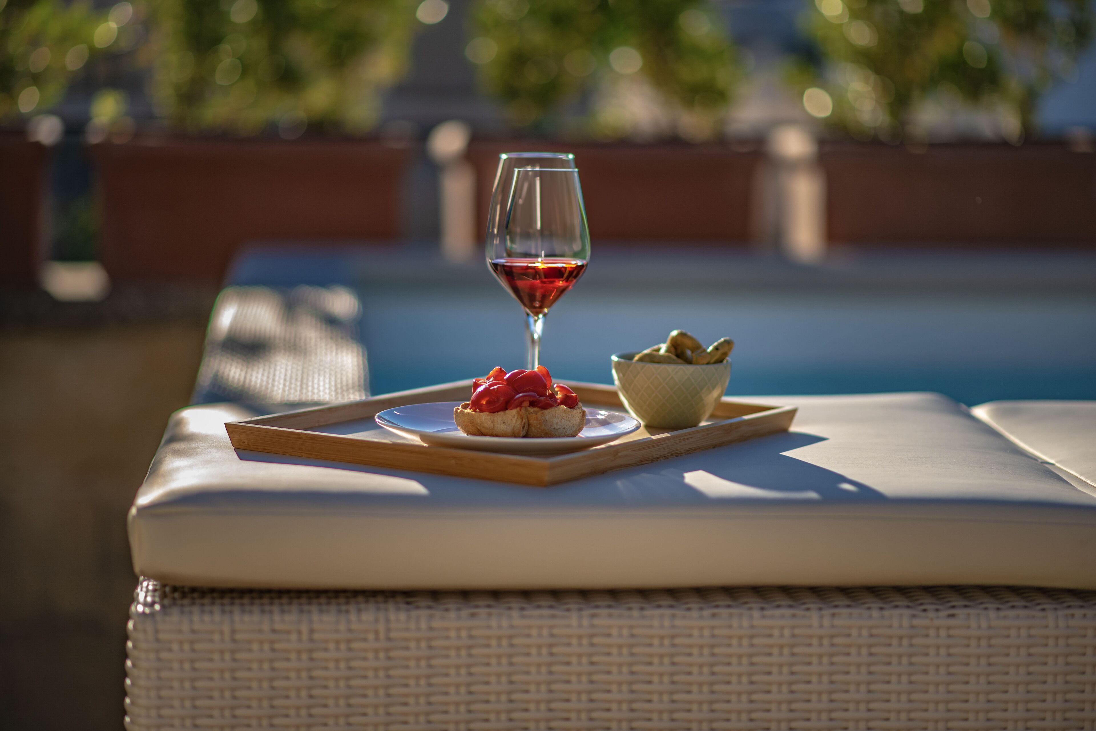 Una bandeja con una copa de vino tinto, un plato pequeño con comida y un cuenco de frutos secos sobre una superficie acolchada cerca de una piscina.