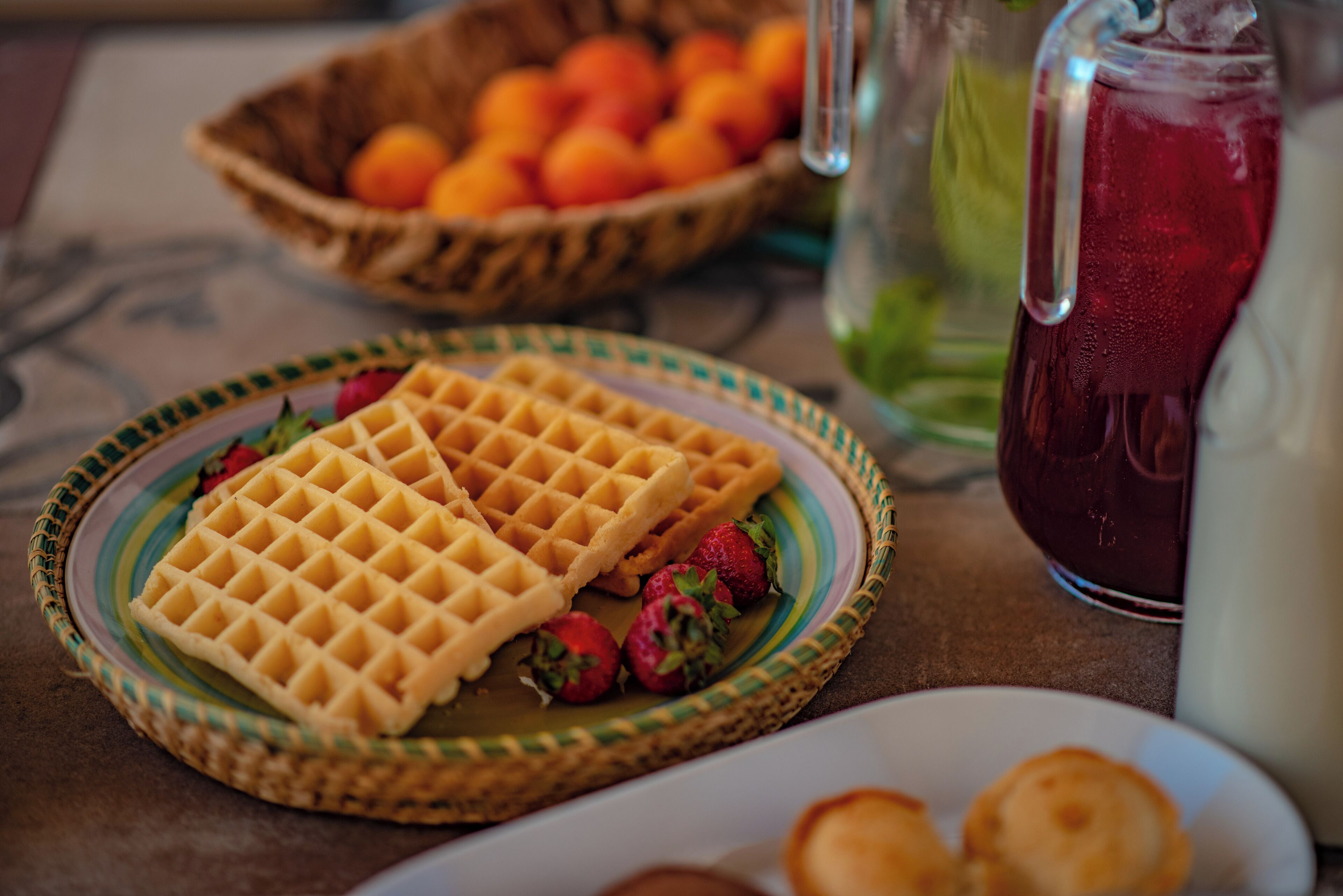 A waffle on a plate with strawberries, a pitcher of red drink, and a basket of oranges.