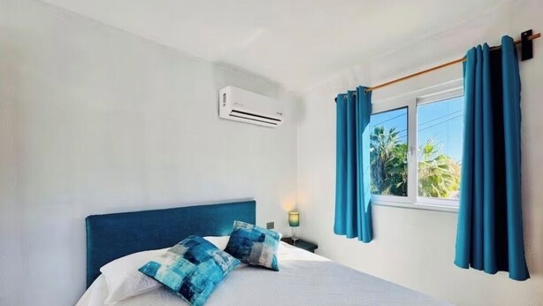 A bedroom with a bed, blue curtains, a window, and an air conditioning unit.
