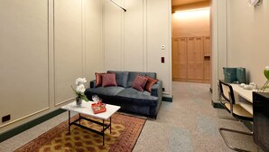 Una sala moderna con un sofá, una mesa de centro y un escritorio pequeño con una silla. Hay una puerta que da a otra habitación y una lámpara fijada a la pared.