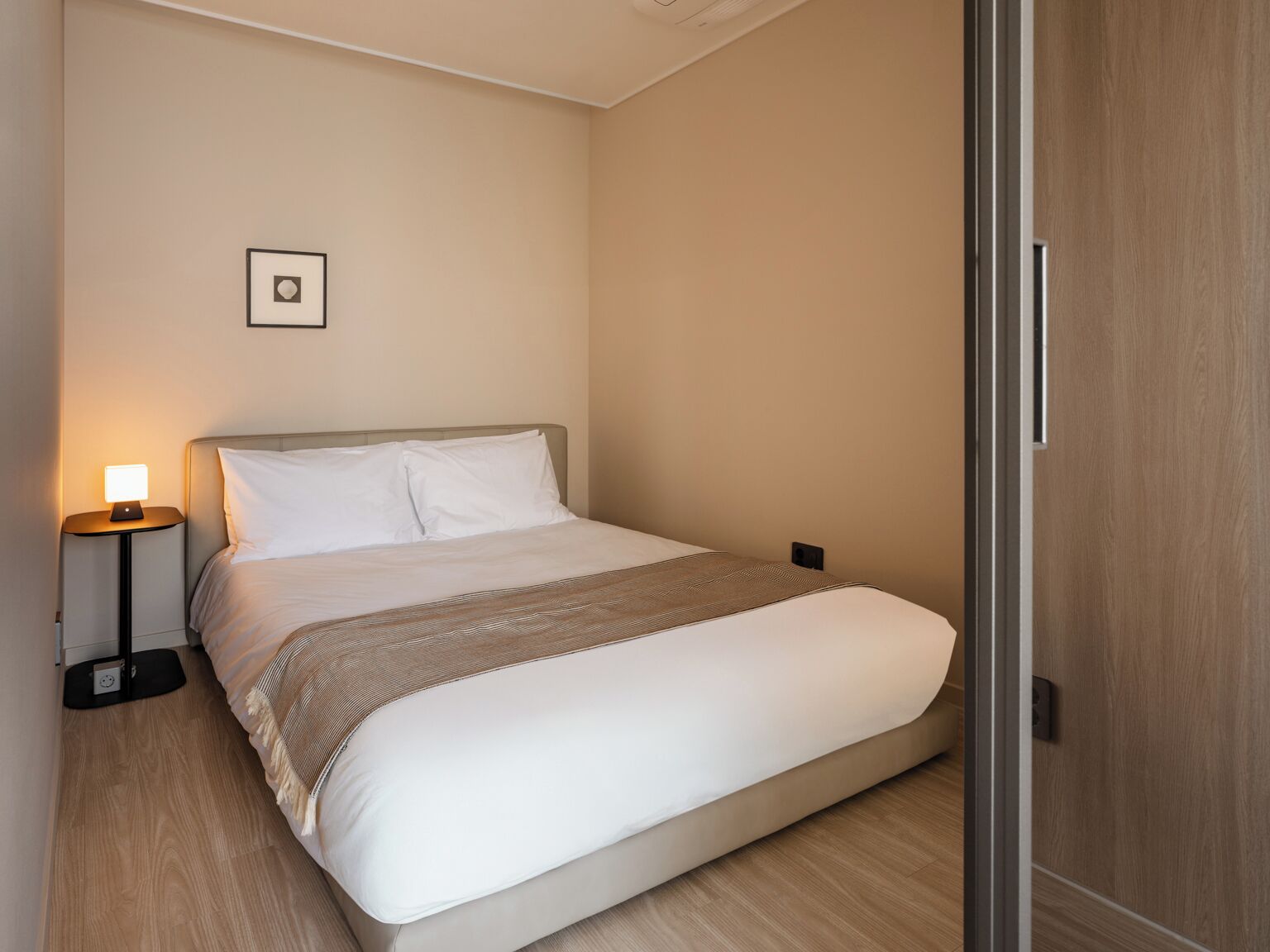 Una cama bien hecha con edredón blanco y manta decorativa estampada, una lámpara de noche y un pequeño cuadro en la pared.