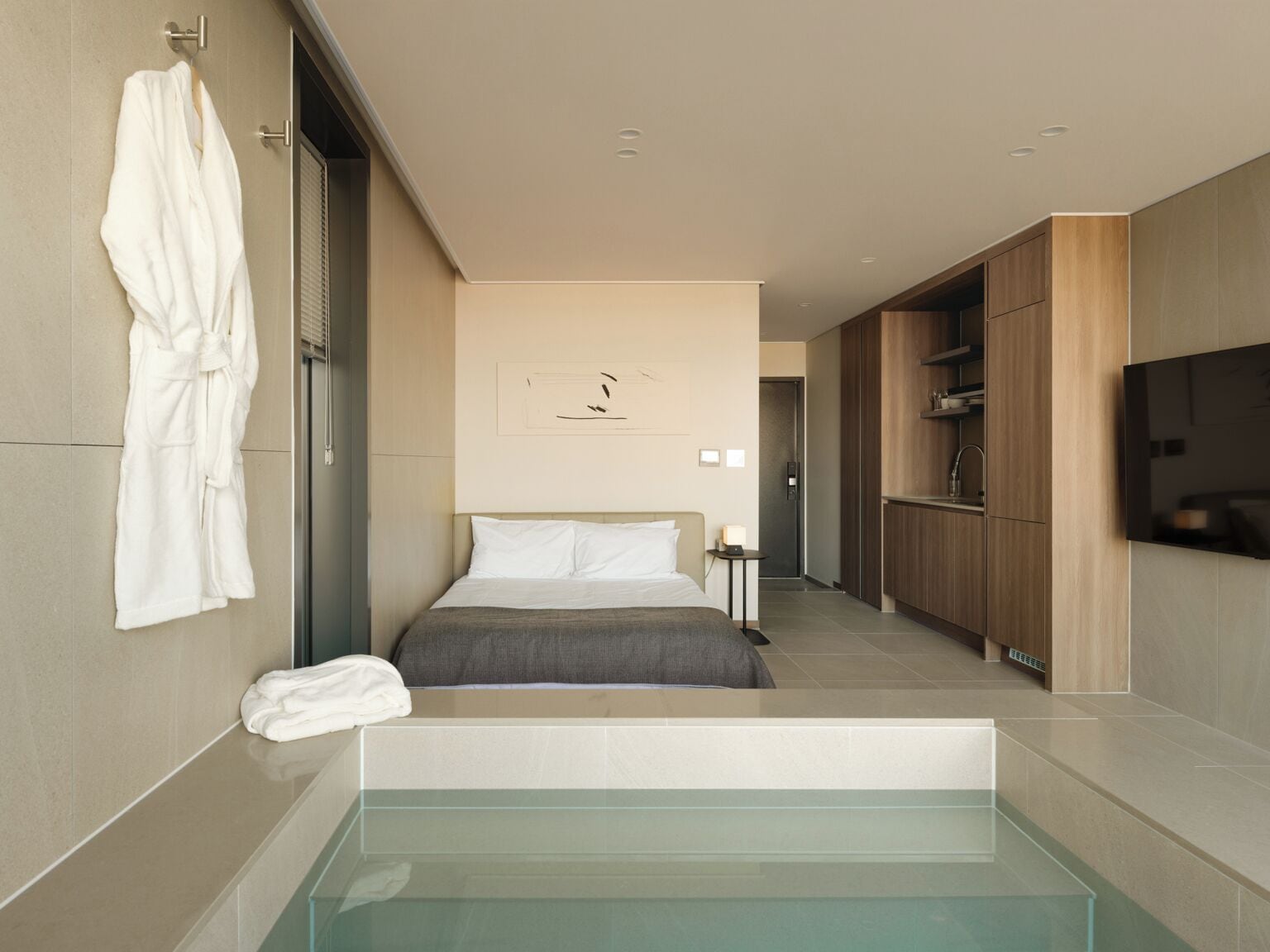 A modern hotel room with a large bed, a glass-enclosed jacuzzi, a wooden wardrobe, and a flat-screen TV mounted on the wall.