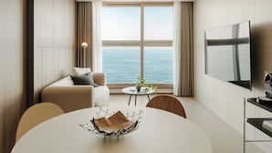 A modern hotel room with a large window offering a view of the sea, a flat-screen TV mounted on the wall, a round dining table with a decorative bowl, and a cozy seating area with a sofa and chairs. - Plott The One Sokcho Xiella The Beach (Sokcho)