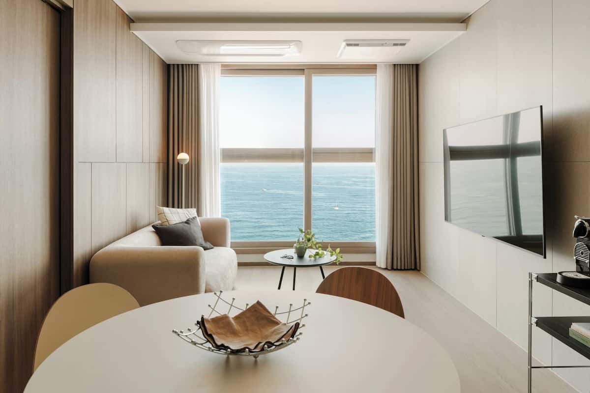 Une chambre d’hôtel moderne dotée d’une grande fenêtre donnant sur la mer, d’un téléviseur à écran plat fixé au mur, d’une table à manger ronde agrémentée d’un vase décoratif, et d’un coin salon confortable avec un canapé et des fauteuils.