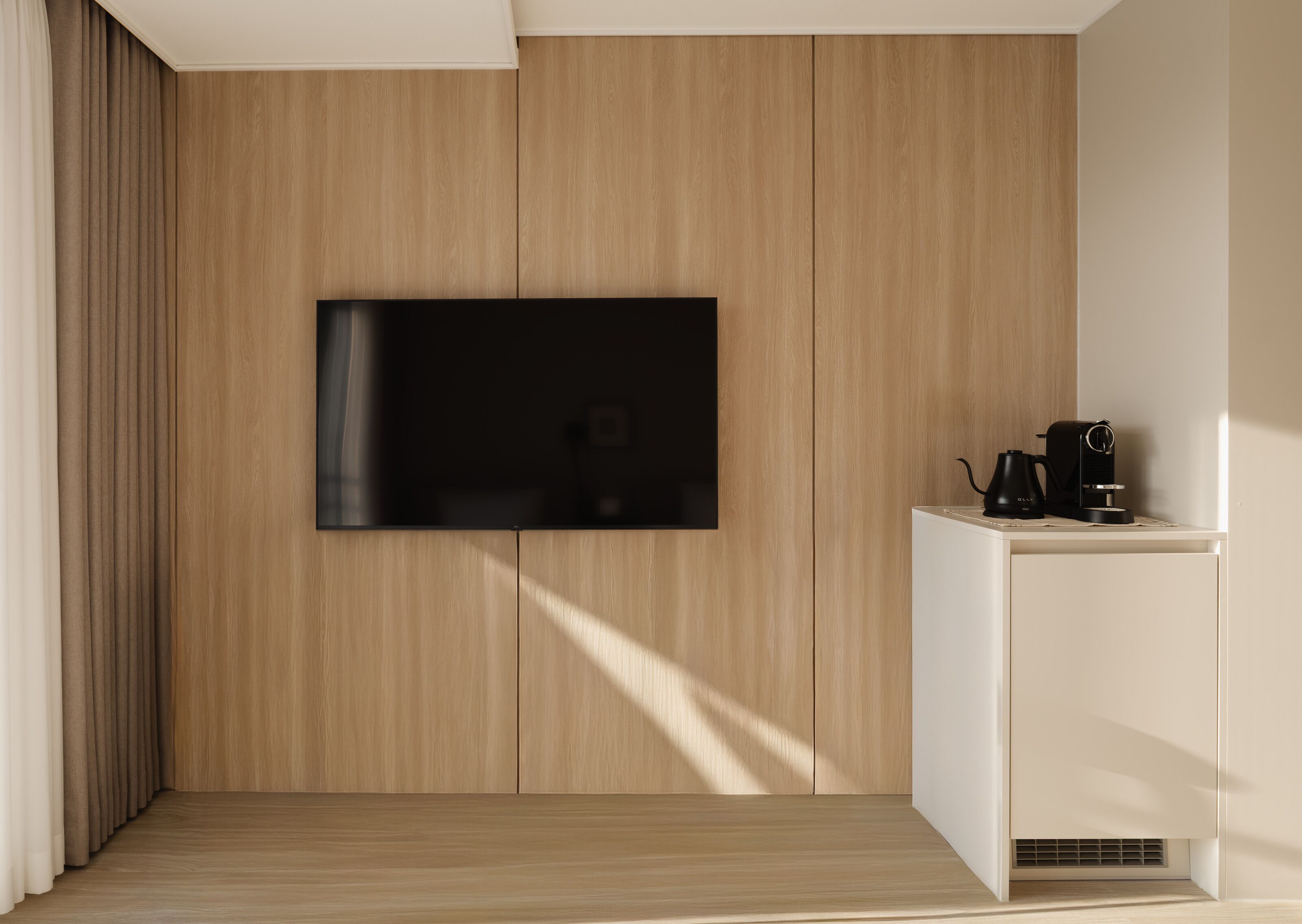 A modern living room with a flat-screen TV mounted on a wooden wall, a coffee maker on a white side table, and a wooden floor.