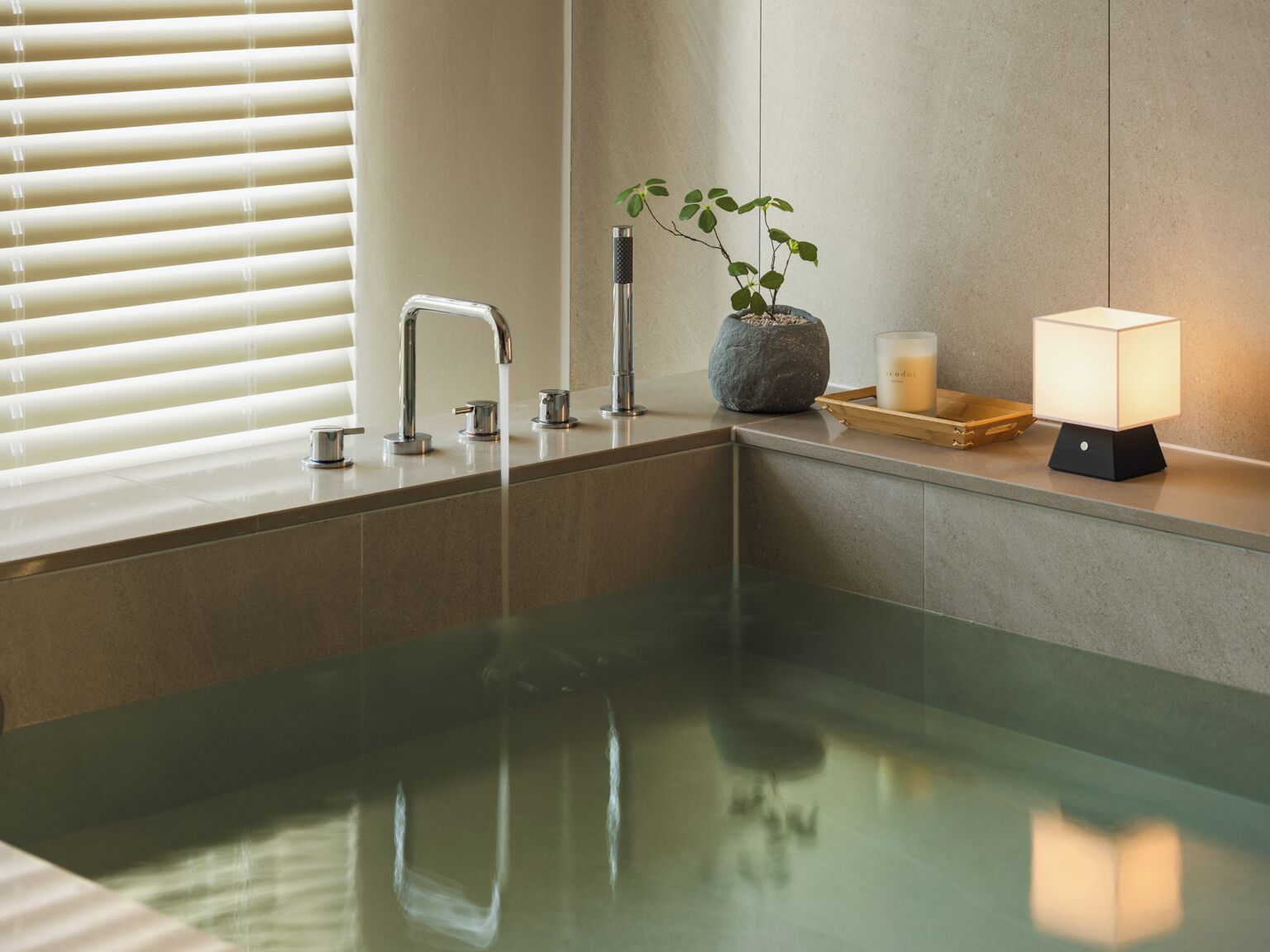A modern bathroom with a glass-enclosed bathtub, a stone planter with a plant, a candle, and a nightstand with a lamp.