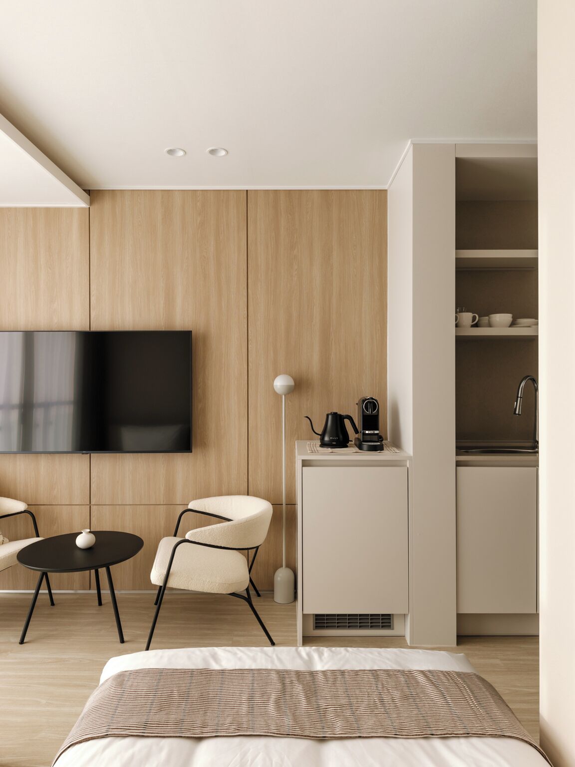 A modern hotel room with a bed, a TV mounted on the wall, a small kitchenette, and a seating area with a table and chairs.