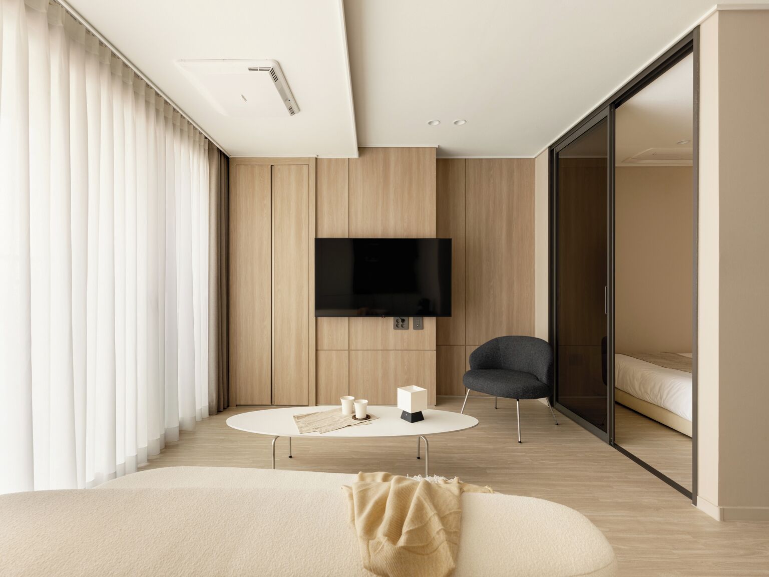A modern hotel room with a large flat-screen TV mounted on the wall, a round coffee table with two candles, a comfortable armchair, and a bed with a sliding glass door leading to a balcony.