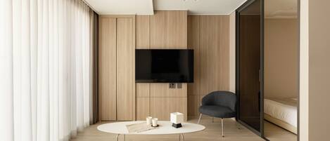 A modern hotel room with a large flat-screen TV mounted on the wall, a round coffee table with two candles, a comfortable armchair, and a bed with a sliding glass door leading to a balcony.