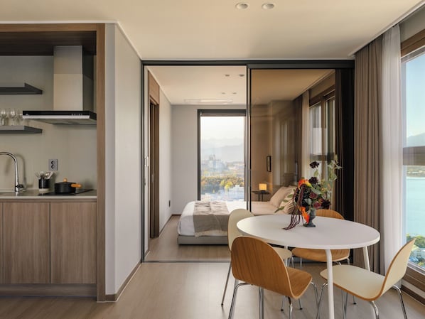 A modern hotel room with a kitchen, a dining area with a round table and chairs, a sofa, and a view of a cityscape through large windows. - Plott The One Sokcho Xiella The Beach (Sokcho)