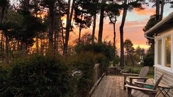 Una terraza de madera con dos sillas, rodeada de vegetación densa y árboles altos, con vistas a un atardecer.