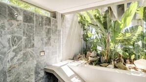 Un baño con pared de piedra, ducha y un ventanal con plantas tropicales.