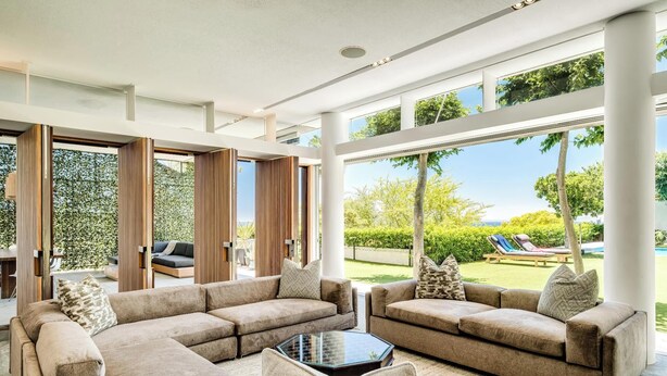 Un amplio salón con un sofá modular grande, una mesa de centro de vidrio y vistas a un jardín exuberante y una piscina.