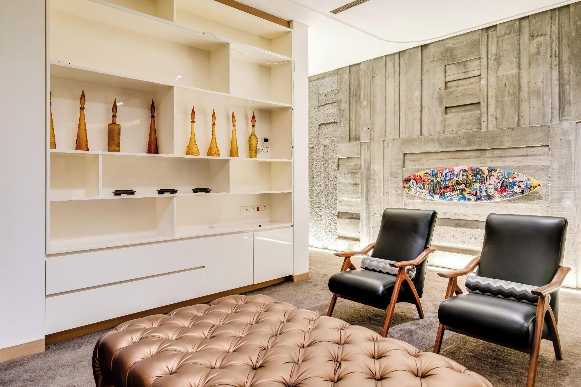 A modern interior with a white shelving unit displaying decorative items, a tufted leather ottoman, and two leather armchairs. A concrete wall with a colorful graffiti mural is visible in the background.