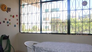 A room with a table and chairs, a window with a metal grid, and a green garden hose on the floor. - The Shell House (Manta Rota)