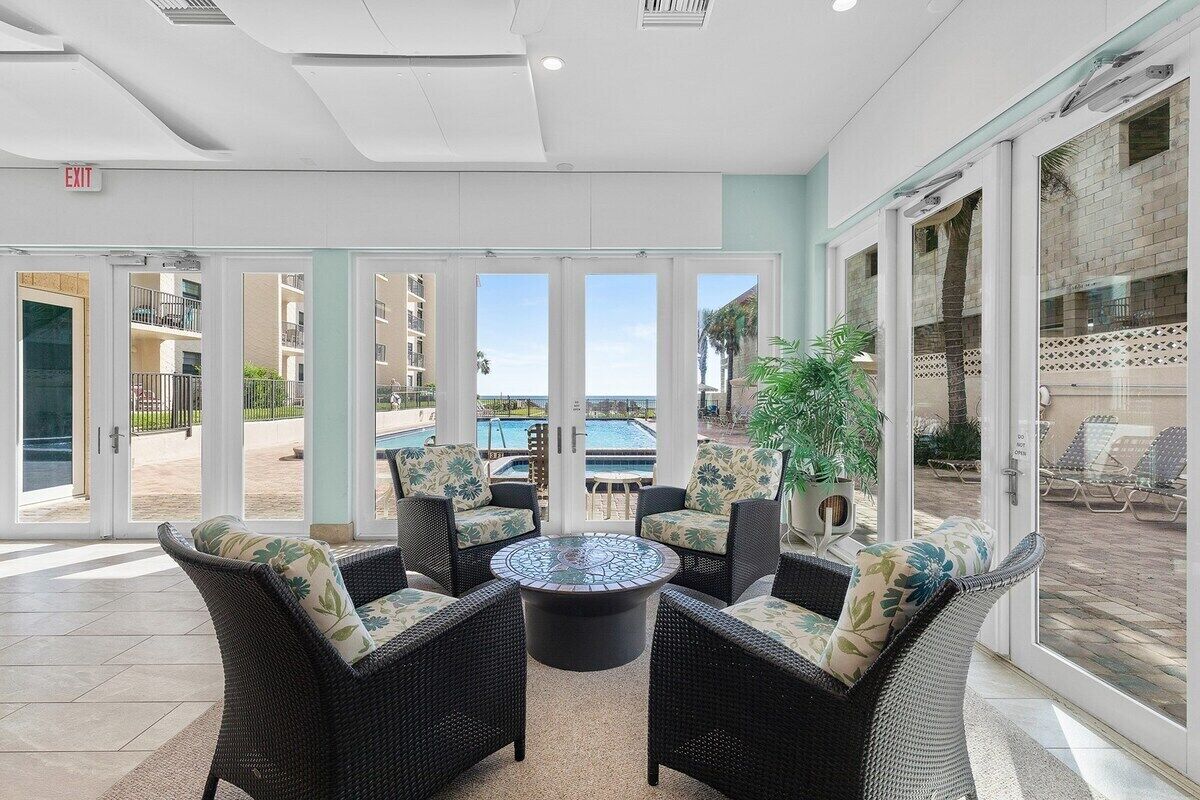 A sunlit indoor seating area with wicker furniture and a central glass-top table, overlooking a pool and outdoor seating.