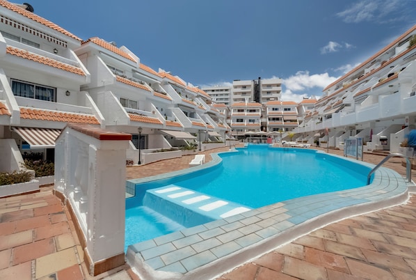 A swimming pool surrounded by white buildings with orange-tiled roofs, a clear blue sky, and a tiled walkway. - Vacation Apartment "Golden Sun Penthouse" with Shared Pool, Private Terrace & Wi-Fi (Playa de las Américas)