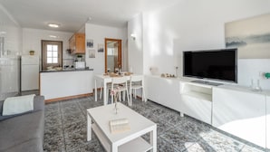 Een moderne keuken met witte kasten, een eethoek met een witte tafel en stoelen, een grijze bank, een flatscreen televisie en een betegelde vloer.