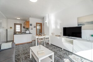 Eine moderne Küche mit weißen Schränken, ein Essbereich mit weißem Tisch und Stühlen, ein graues Sofa, ein Flachbildfernseher und ein gefliester Boden.