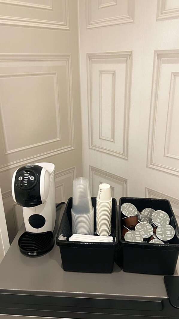 Una cafetera, vasos de plástico apilados y un recipiente con tapas sobre una mesa junto a una puerta.