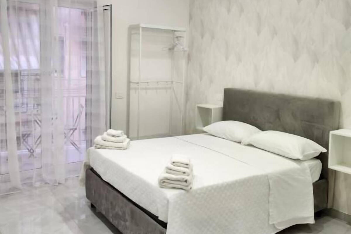 A modern bedroom with a bed, white linens, a grey headboard, a bedside table, and a bathroom with a shower and toilet.