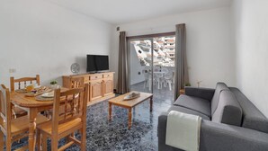 Ein Wohnzimmer mit einer Couch, einem Holzesstisch, Stühlen, einem Fernseher und einem Balkon mit Sitzgelegenheiten.