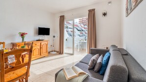 A living room with a sofa, a dining table, a TV, and a balcony with curtains. - Apartment Infinity Pool Views with Shared Pool, Private Terrace and Wi-Fi (Playa de las Américas)