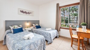 A hotel room with two beds, a nightstand, a table, and a window with a view of palm trees. - Apartment Infinity Pool Views with Shared Pool, Private Terrace and Wi-Fi (Playa de las Américas)