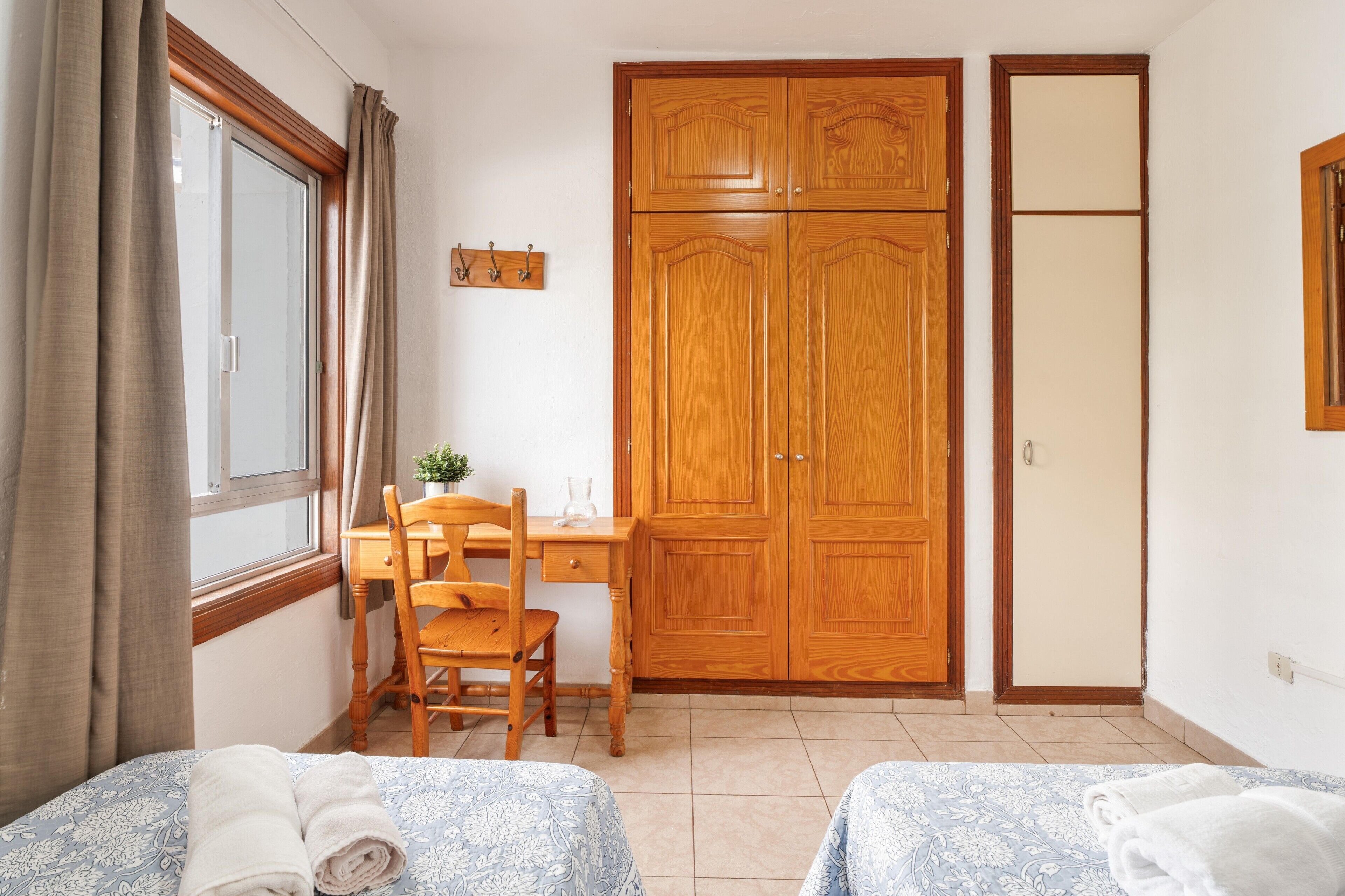 A bedroom with two beds, a wooden wardrobe, a desk with a chair, and a window with curtains.