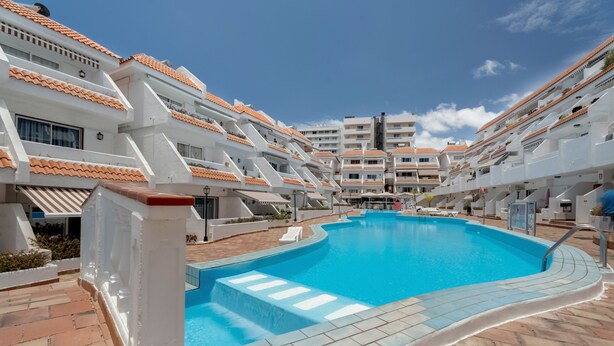 A swimming pool surrounded by white buildings with orange-tiled roofs, a clear blue sky, and a tiled walkway. - Apartment Infinity Pool Views with Shared Pool, Private Terrace and Wi-Fi (Playa de las Américas)