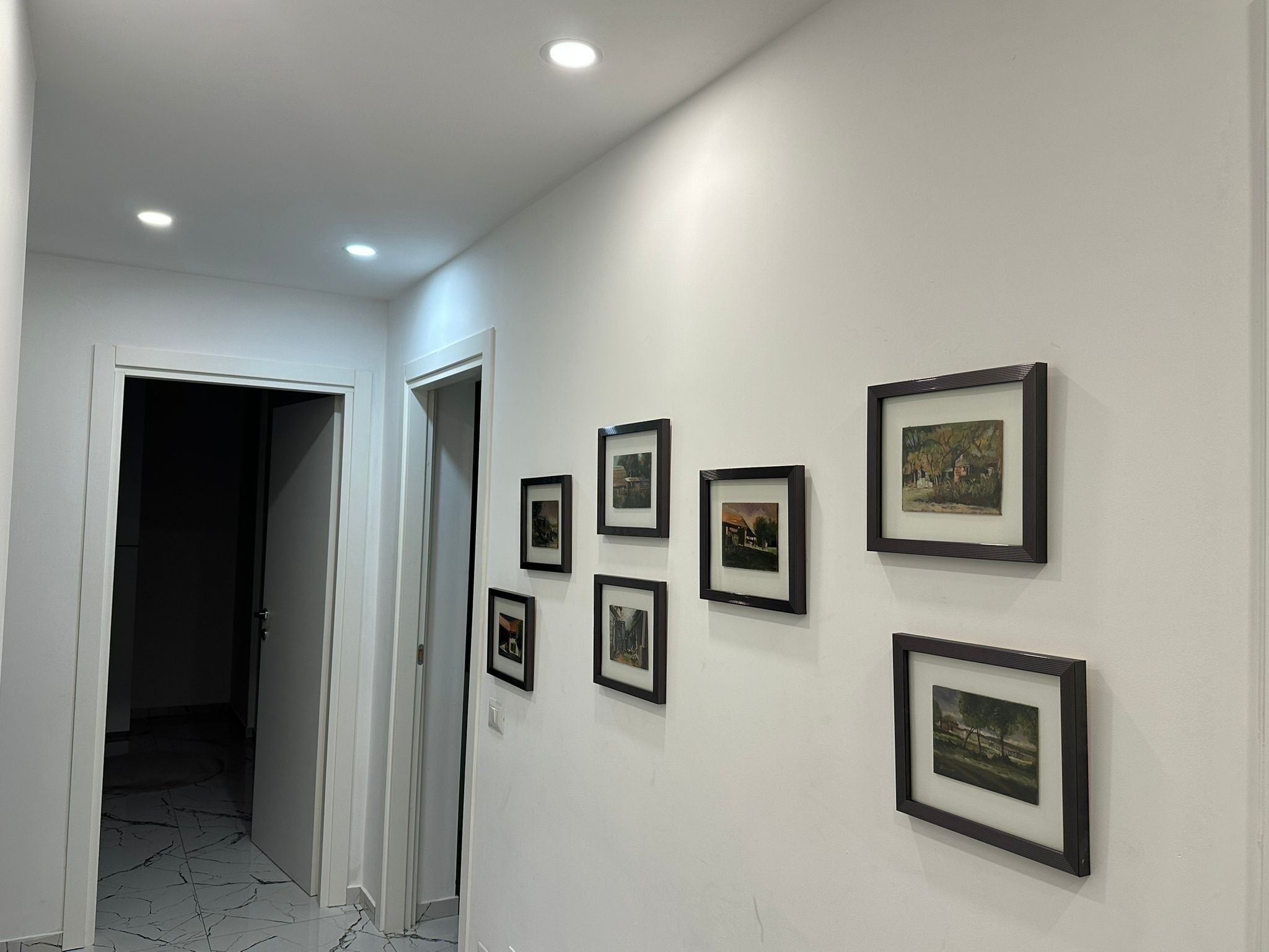 Un couloir aux murs blancs et au sol carrelé. Plusieurs tableaux encadrés sont accrochés au mur.