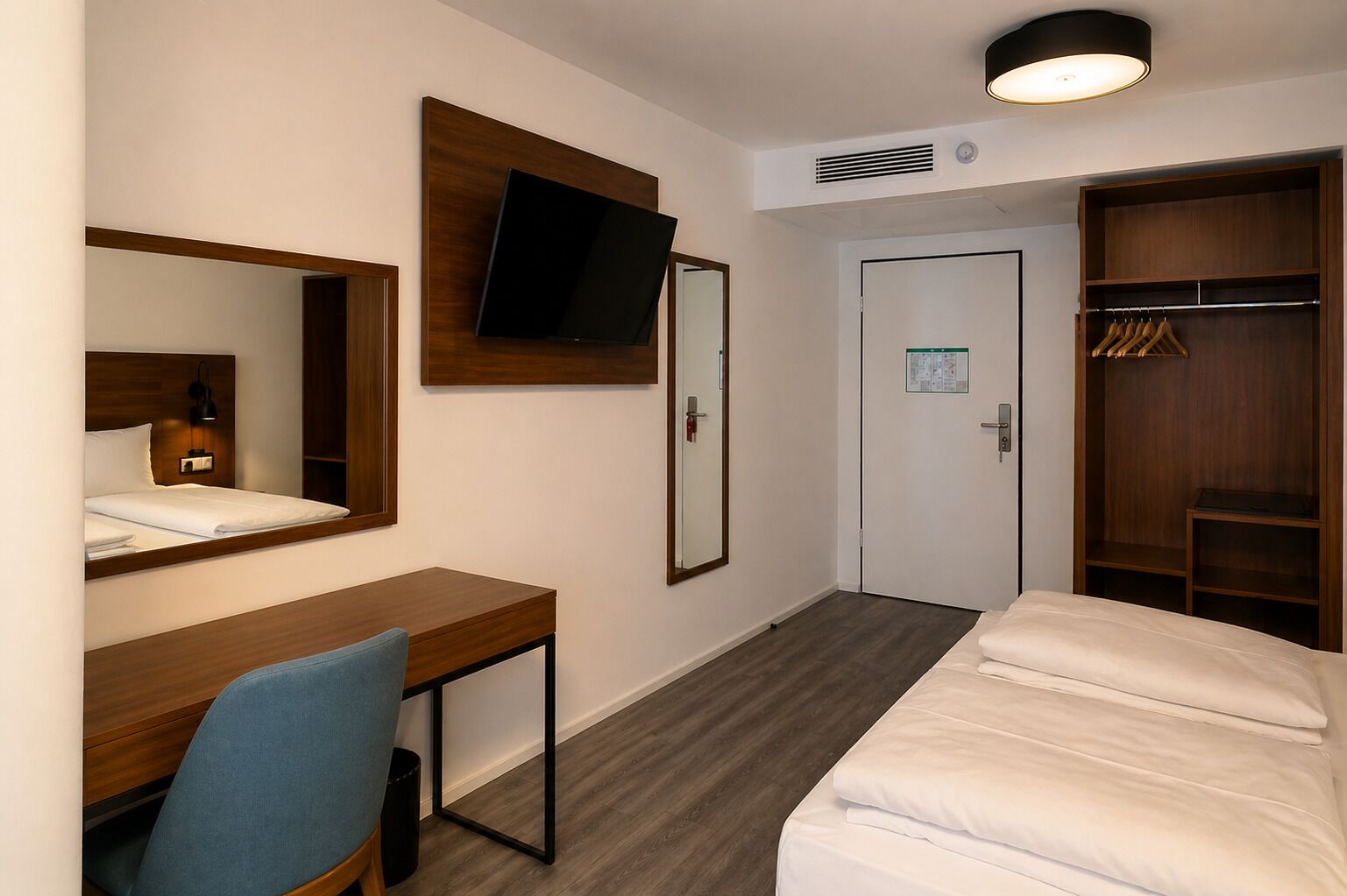Ein Hotelzimmer mit Bett, Schreibtisch, Stuhl, Wandfernseher, Kleiderschrank und einer Tür mit Kalender.