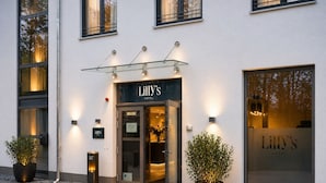A hotel entrance with a glass canopy, a sign reading 'Lilly's Hotel,' and two potted plants on the ground. - Lillys Hotel München-Unterschleißheim  (Unterschleißheim)