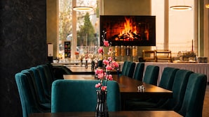 A restaurant with teal chairs, a wooden table, a vase with pink flowers, and a TV displaying a fire. - Lillys Hotel München-Unterschleißheim  (Unterschleißheim)