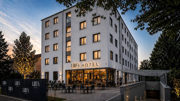 A multi-story hotel with a prominent entrance, outdoor seating area, and a covered walkway. - Lillys Hotel München-Unterschleißheim  (Unterschleißheim)