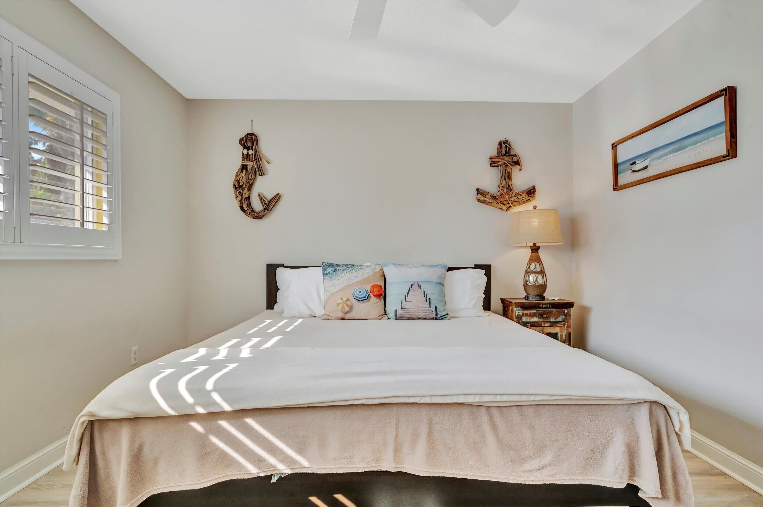 A neatly made bed with a beige cover, a nightstand with a lamp, and wall decorations including a framed picture of a beach scene.