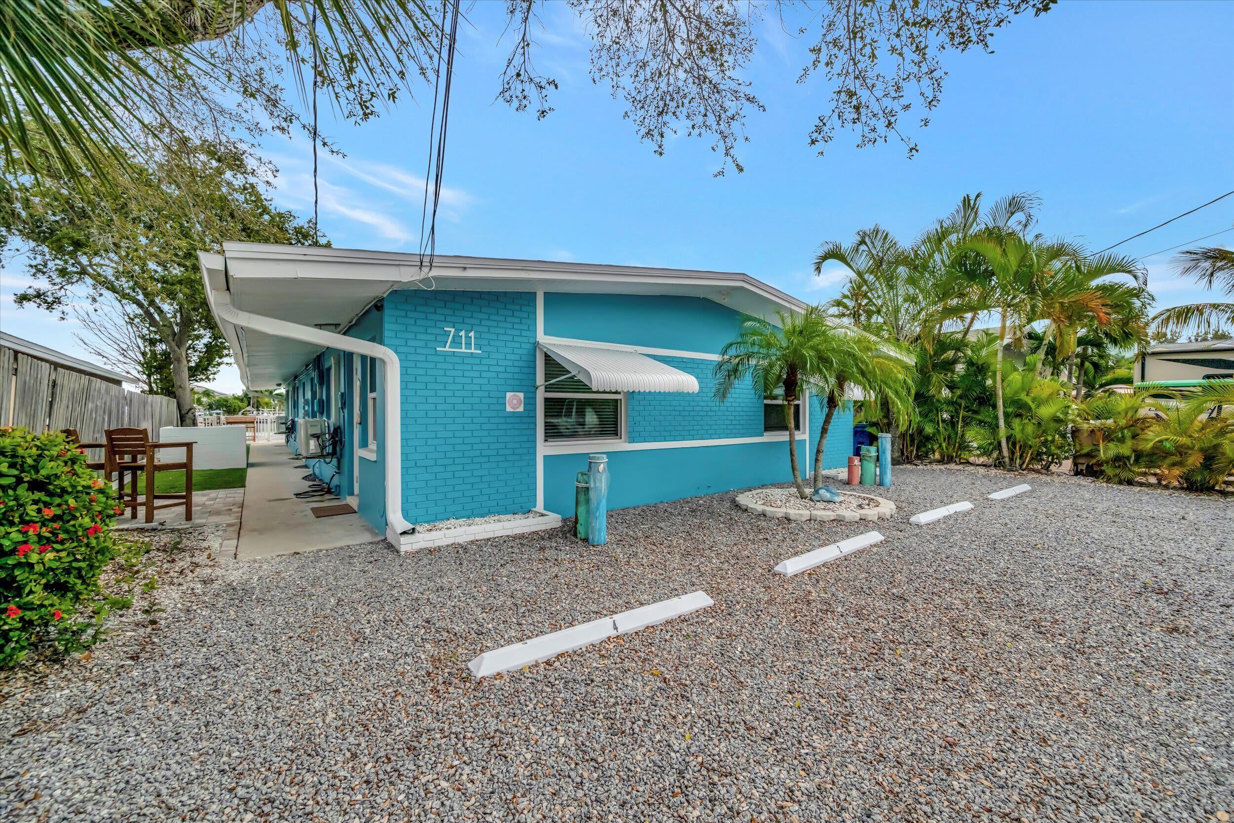 A turquoise building with a white awning, a gravel driveway, and a palm tree in the background.