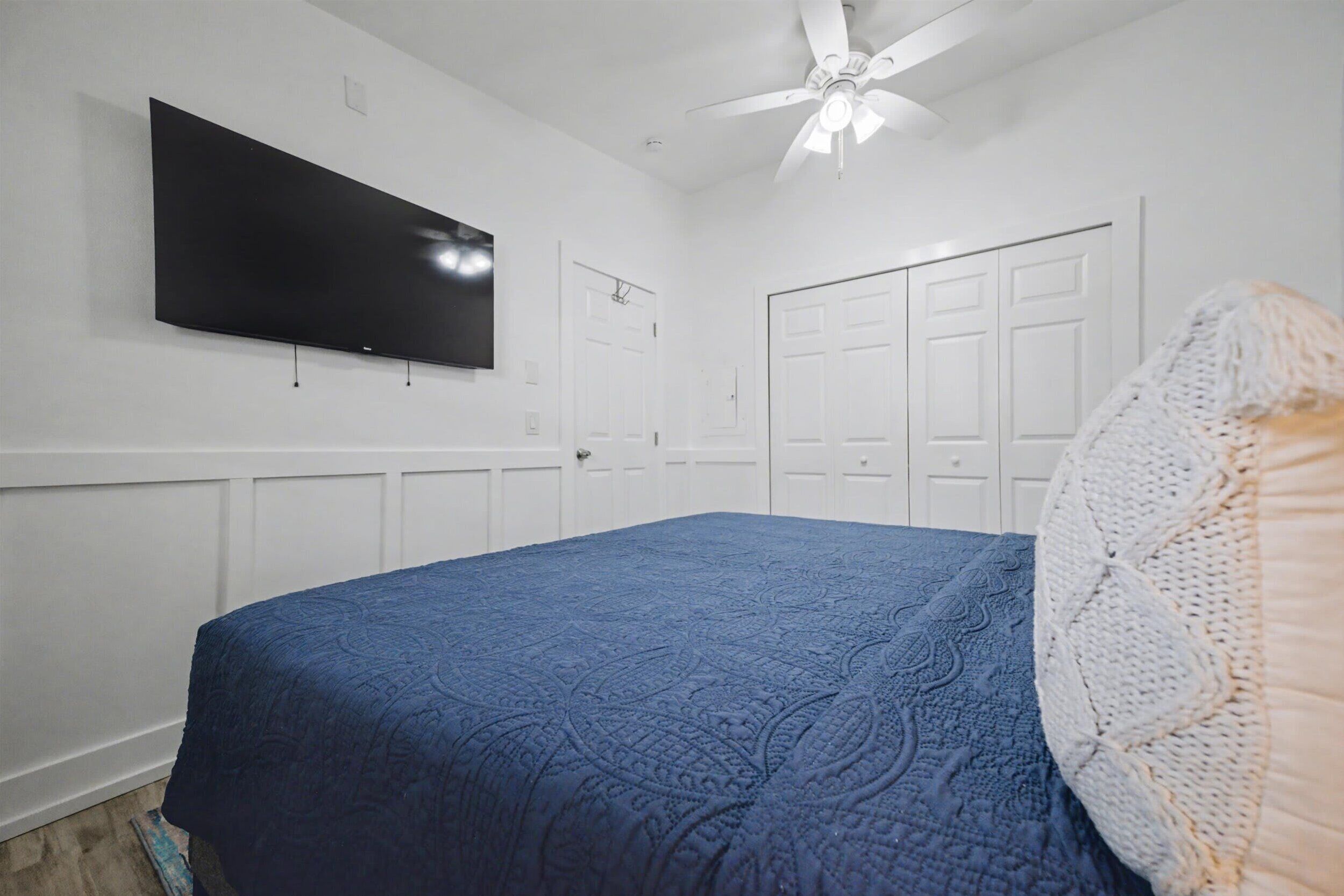 A bedroom with a large bed, a ceiling fan, and a flat-screen TV mounted on the wall.