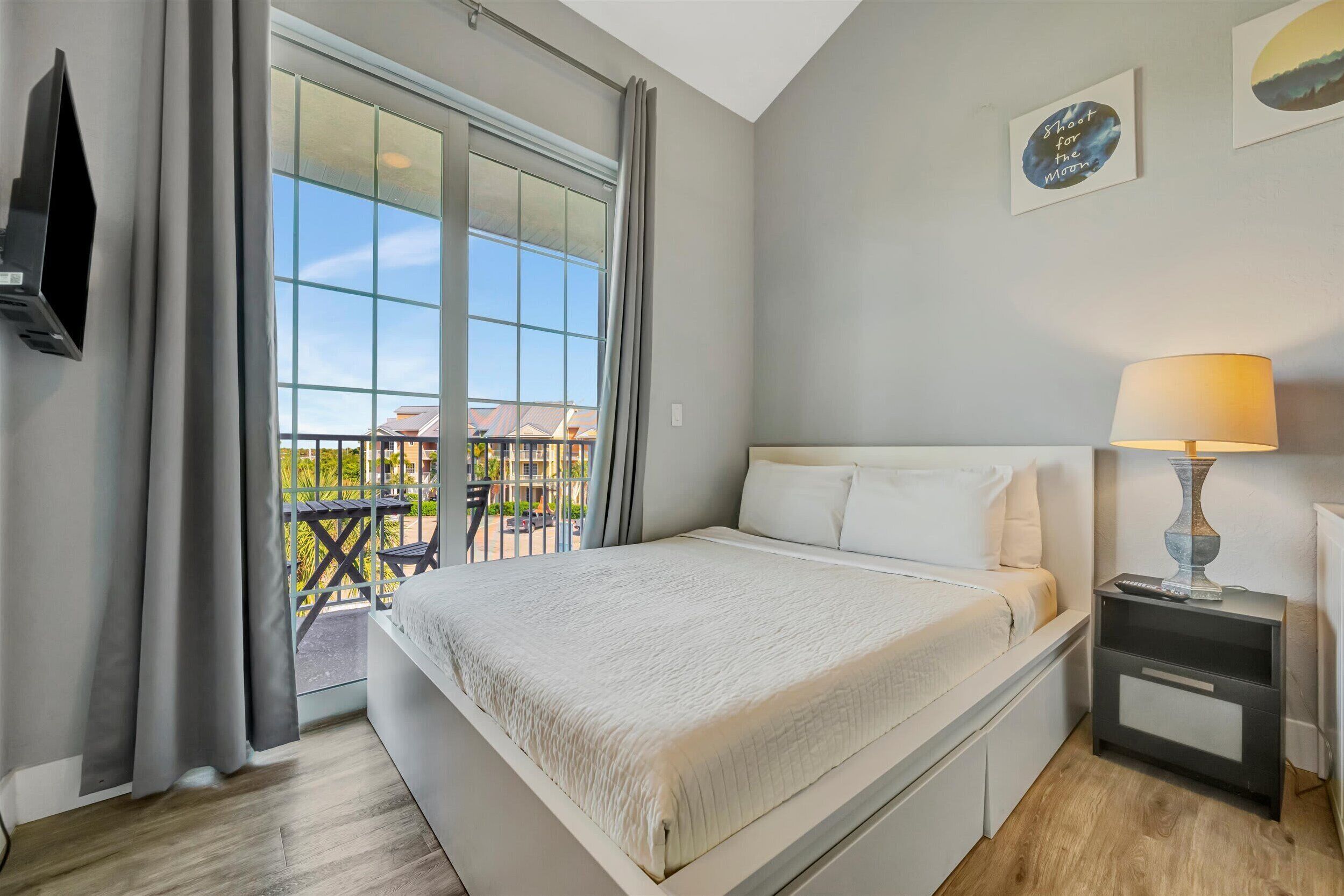 A bedroom with a large bed, a nightstand with a lamp, a TV mounted on the wall, and a balcony with a view of buildings.